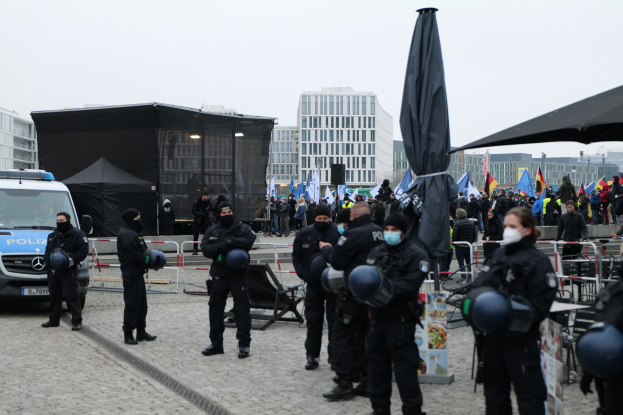 Eine Gruppe von Polizeibeamten steht vor einer Menge von Demonstranten in Berlin, Deutschland, mit Barrikaden, Zelten und Fahnen im Vordergrund und Gebäuden im Hintergrund unter einem klaren Himmel.