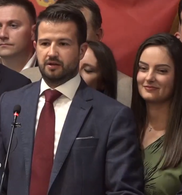 Ein Mann in Anzug und Krawatte steht an einem Rednerpult und spricht zu einer Menge mit einem Banner im Hintergrund, wahrscheinlich zu einem Regierungsverbot von Social Media.