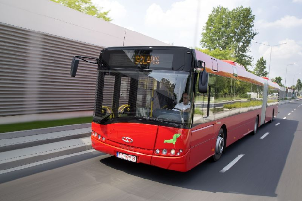 Ein roter Bus fährt auf einer Straße an einem Gebäude vorbei, mit einer Person im Inneren, während sich auf der linken Seite eine Wand befindet und im Hintergrund Bäume, Laternenpfähle und ein bewölkter Himmel zu sehen sind.