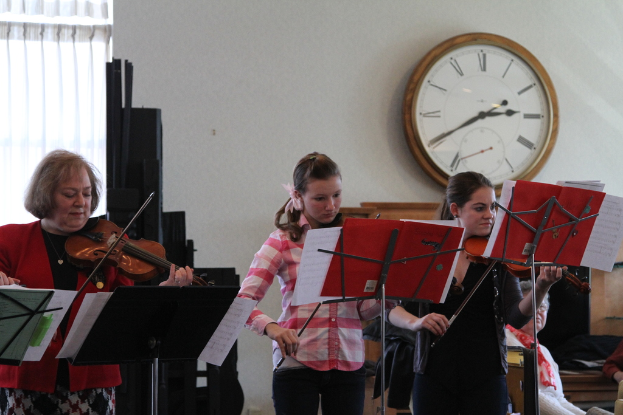 Drei Frauen auf der Bühne mit Geigen stehend und auf Musikständern spielend, während eine Gruppe von sitzenden Frauen im Vordergrund zuhört; eine Uhr hängt an der weißen Wand im Hintergrund, mit einem weißen Vorhang in der linken Ecke.