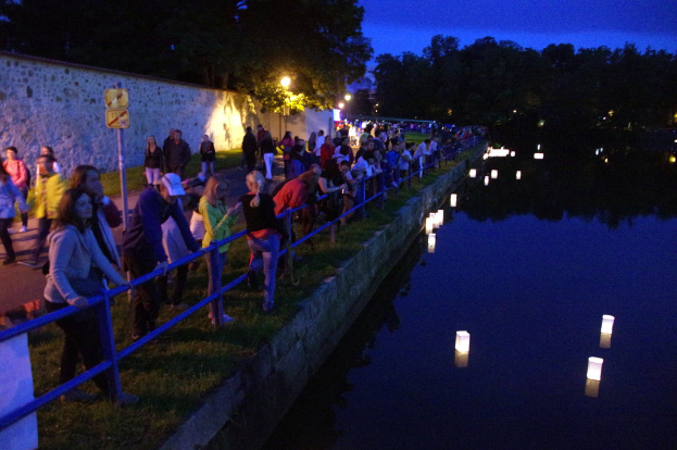 Ein Kasten voller Kerzen, die auf dem Wasser schwimmen, mit Menschen, die auf dem Rasen und auf der Straße stehen und gehen, umgeben von Bäumen, Pfählen und Lichtern unter dem Himmel.