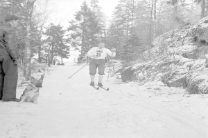 Schwarzes und weißes Foto eines Mannes, der auf Skiern steht und Stöcke hält, begleitet von einem Hund, der neben ihm sitzt, mit Bäumen und einem klaren Himmel im Hintergrund.