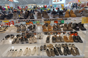 Ein Schuhgeschäftsinneres mit verschiedenen Schuharten auf Gestellen, Preisetiketten sichtbar, Menschen im Hintergrund, Kleidung an Bügeln, Deckenlampen, ein Bildschirm und Informationsschilder.