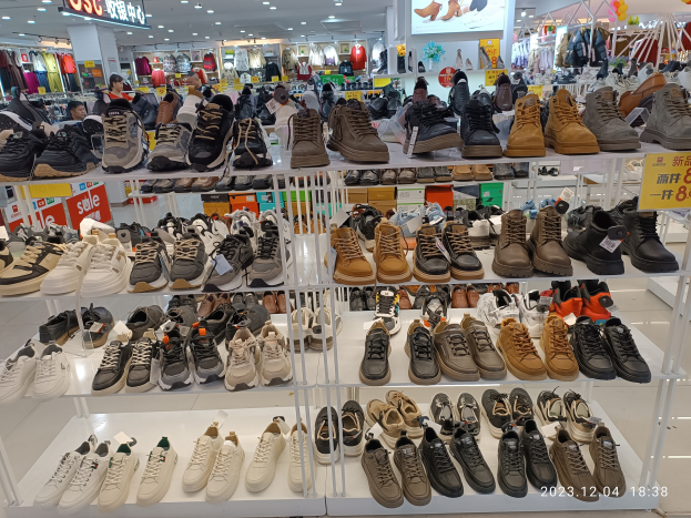 Ein Schuhgeschäftsinneres mit verschiedenen Schuharten auf Gestellen, Preisetiketten sichtbar, Menschen im Hintergrund, Kleidung an Bügeln, Deckenlampen, ein Bildschirm und Informationsschilder.