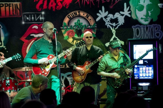 Drei Männer spielen Gitarren auf einer Bühne bei einem Konzert, mit einer Menge darunter und einer gemalten Wand im Hintergrund.