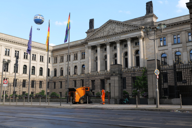 Großes modernes Gebäude mit zahlreichen Fenstern, als Bundestag in Berlin, Deutschland identifiziert, umgeben von Straßeninfrastruktur, Fahrzeugen, Fußgängern, Flaggen und Bäumen unter einem bewölkten Himmel.