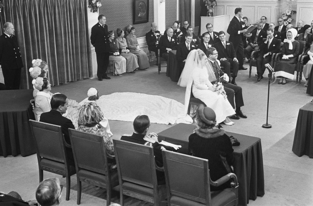 Schwarzes und weißes Foto einer Hochzeitszeremonie mit sitzenden und stehenden Gästen, einem Mann mit Mikrofon, einer Frau mit einem Strauß, einem Podium mit Mikrofon, Blumenschmuck, einem Vorhang, einem gerahmten Foto an der Wand und einer Tür im Hintergrund.