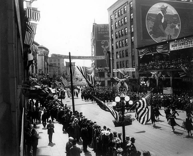Eine große Menge marschiert auf einer Stadtstraße während eines Umzugs, hält Fahnen mit Gebäuden und Laternenmasten an den Seiten, in Schwarz-Weiß.