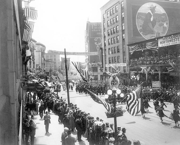 Eine große Menge marschiert auf einer Stadtstraße während eines Umzugs, hält Fahnen mit Gebäuden und Laternenmasten an den Seiten, in Schwarz-Weiß.