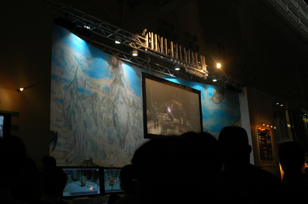 Eine Bühne mit einem Video-Display in der Mitte, vor der eine Gruppe von Menschen sitzt.