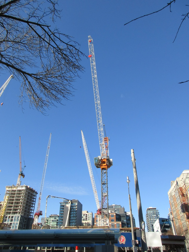 Eine Gruppe von Baukränen auf einer Stadtstraße umgeben von Gebäuden, Bäumen, Polen, Brettern und anderen Gegenständen mit dem Himmel im Hintergrund.