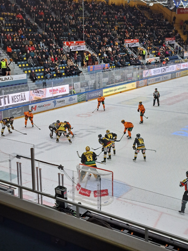Eine Gruppe von Menschen, die Hockey auf einer Indoor-Eisfläche spielen, umgeben von einem Zaun, mit Zuschauern im Hintergrund und sichtbaren Bannern.
