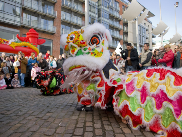 Ein farbenfrohes chinesisches Neujahrsfest in Amsterdam mit einem Löwen Tanz und einer Zuschauermenge, einige machen Fotos, vor einer Kulisse aus Gebäuden, Laternenmasten und einem klaren blauen Himmel.