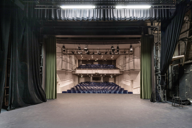 Großer Zuschauerraum mit schwarzen Vorhängen, einer beleuchteten Bühne und Sitzreihen, mit Gegenständen auf der rechten Seite, die auf eine Veranstaltungslocation hinweisen.