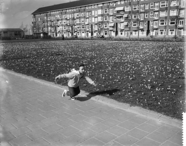 Ein junger Junge rennt über einen Gehweg in einem Park, umgeben von Gebäuden, Bäumen und einem klaren Himmel, trägt Schuhe.