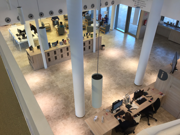 Großraumbüro mit Menschen an Schreibtischen mit Monitoren, Tastaturen und anderen Büromaterialien, mit Deckenleuchten und Säulen, einem sichtbaren Feuerlöscher und einer Tafel mit Text auf der rechten Seite.