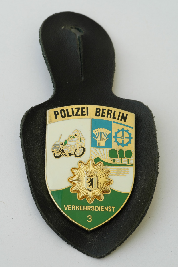 Ein schildförmiges Polizeibadgebnis mit "Polizei Berlin" in schwarzer Fettschrift, das ein weißes Stern auf einem blauen Hintergrund und einen weißen Rand hat und auf einer Oberfläche ruht.