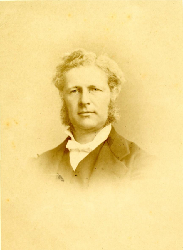 Ein altes Foto eines Mannes mit ordentlich gekämmten Locken, der einen maßgeschneiderten Anzug trägt und direkt in die Kamera schaut.