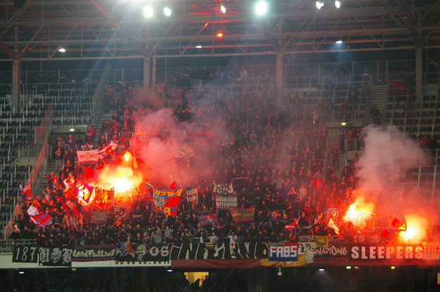 Eine große Menge Menschen in einem Stadion hält Fahnen und Banner, mit Leuchtraketen, die Rauch erzeugen, unter einer Decke mit Lampen und Metallrahmen.