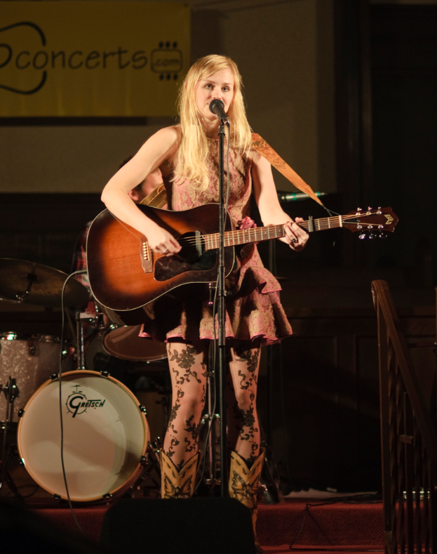 Eine Frau spielt Gitarre und singt auf der Bühne bei einem Konzert, mit einer Band im Hintergrund und einem Bildschirm an der Wand dahinter.