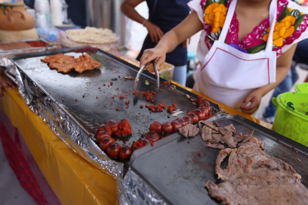 Eine Frau in einer Schürze grillt Essen auf einem Markt, mit einem Tisch voller Lebensmittel, einem Eimer und anderen Gegenständen in der Nähe und ein paar Menschen im Hintergrund.
