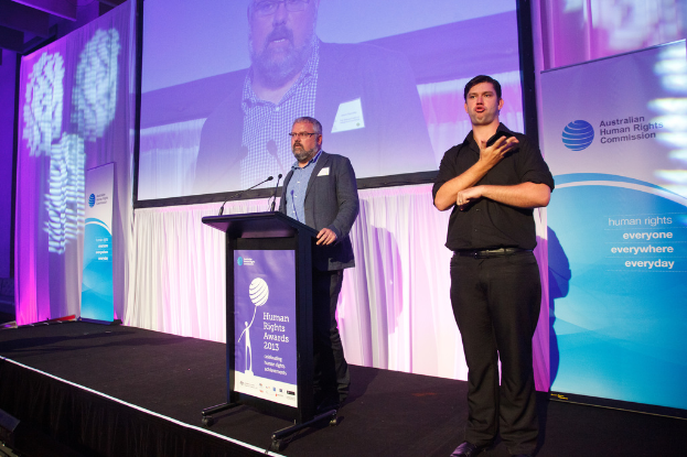 Eine Person in einem schwarzen Kleid steht rechts neben einem Konferenzsaal, während ein Mann in einem schwarzen Anzug auf einem Podium mit drei Mikrofonen in der Mitte spricht, flankiert von Werbetafeln auf beiden Seiten, mit einem Vorhang und einem Bildschirm im Hintergrund.