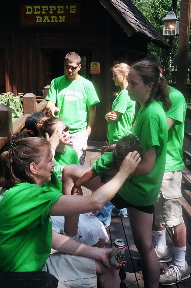 Mehrere Personen in grünen T-Shirts, einige sitzend und einige stehend, vor einer hölzernen Hütte, Bäumen und einer Lampe, mit einem hölzernen Zaun und Pflanzen im Hintergrund.