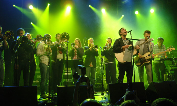 Eine Gruppe von Musikern, die auf der Bühne mit Mikrofonen, Scheinwerfern und einer Kamera spielen, vor einem Publikum.