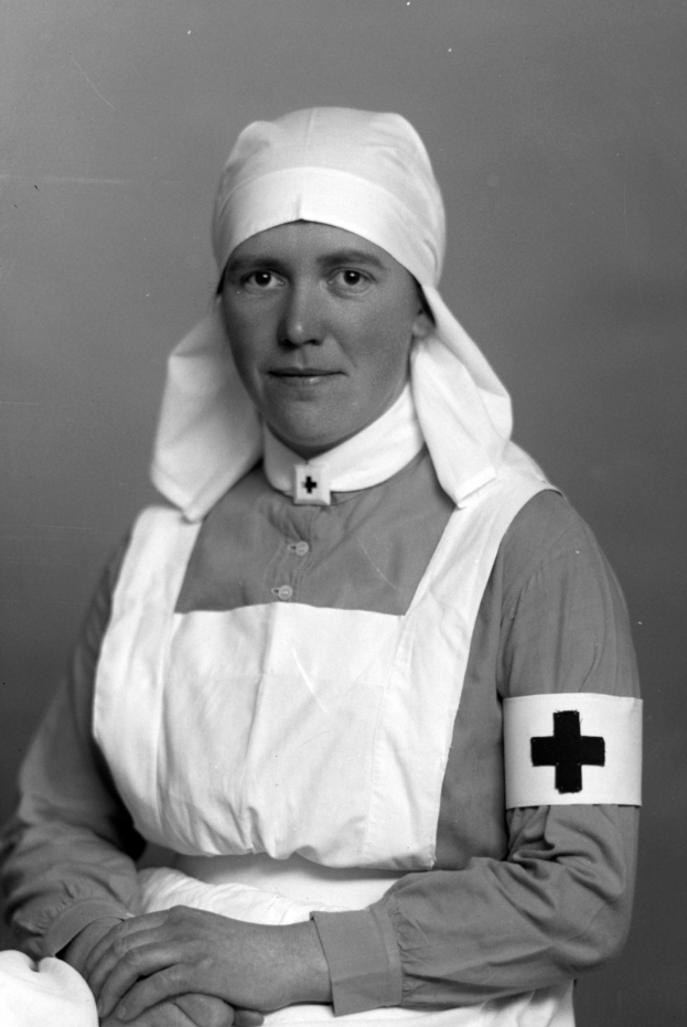 Ein Schwarz-Weiß-Foto einer Frau in einer Krankenschwester-Uniform, die einen weißen Kittel mit einem roten Kreuz trägt, ein Tuch hält und ihre Haare zu einem Dutt gebunden hat, die direkt in die Kamera schaut.