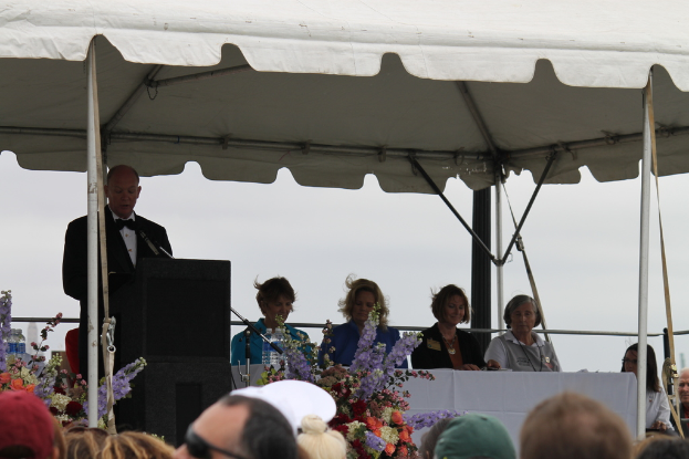 Eine Gruppe von Menschen sitzt an einem Tisch mit Blumen, Flaschen und einem Mikrofon auf einem Ständer, während eine Person in der Nähe eines Podiums unter einem Pavillon mit sichtbaren Pfählen steht.