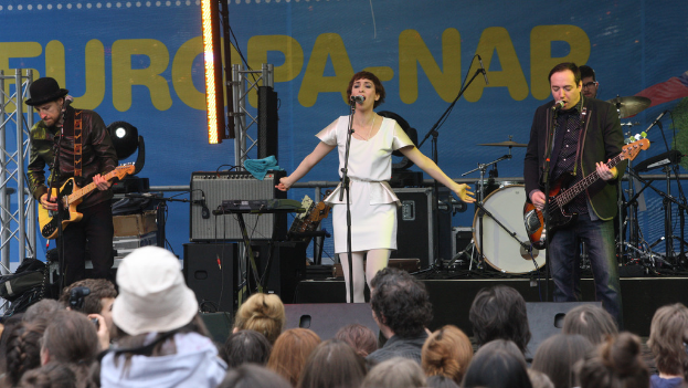 Eine Bühne mit vier Musikern: eine Frau in einem weißen Kleid singt in der Mitte, zwei Männer spielen Gitarren links und rechts und eine Menge ist unten zu sehen mit einer Wand und Lautsprechern im Hintergrund.