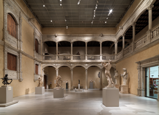 Ein großer Museumsraum mit zahlreichen Statuen und Skulpturen auf Sockeln, architektonischen Elementen wie Wänden, Säulen und Geländern, Deckenleuchten und einigen Besuchern.