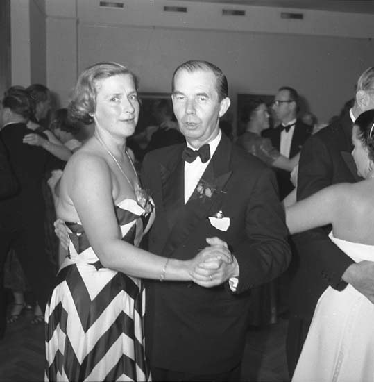 Ein Mann in einem schwarzen Blazer, weißem Hemd und schwarzen Hosen tanzt mit einer Frau in einem unbekannten Outfit auf einer formellen Veranstaltung, indem sie sich an den Händen halten und zur Musik an einer Wand schwenken.