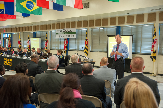 Eine Gruppe von Menschen, die auf Stühlen in einem Raum sitzen, mit einem Redner vor einem Projektionsschirm und einer Tafel mit Wasserflaschen, Flaggenmasten, hängenden Flaggen, abgedeckten Fenstern und einer Tür auf der linken Seite.