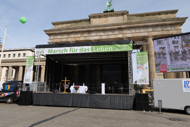 Eine Bühne vor dem Brandenburgertor in Berlin, Deutschland, mit einem Tisch mit einer Blumenvase, einem Kreuzsymbol und anderen Gegenständen sowie Bannern mit Text, Lautsprechern und Fahrzeugen auf der Straße. Im Hintergrund sind Gebäude mit Fenstern, eine Statue auf einem Gebäude, ein Mast mit einer Flagge und ein bewölkter Himmel zu sehen.