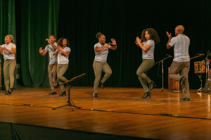 Eine Gruppe von Menschen auf einer Bühne, die klatschen und feiern, mit Mikrofonen, Musikinstrumenten und einem Vorhang im Hintergrund, wahrscheinlich eine Tanzgruppe, die ihren Erfolg anerkennt.