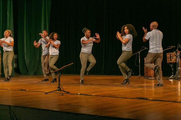 Eine Gruppe von Menschen auf einer Bühne, die klatschen und feiern, mit Mikrofonen, Musikinstrumenten und einem Vorhang im Hintergrund, wahrscheinlich eine Tanzgruppe, die ihren Erfolg anerkennt.