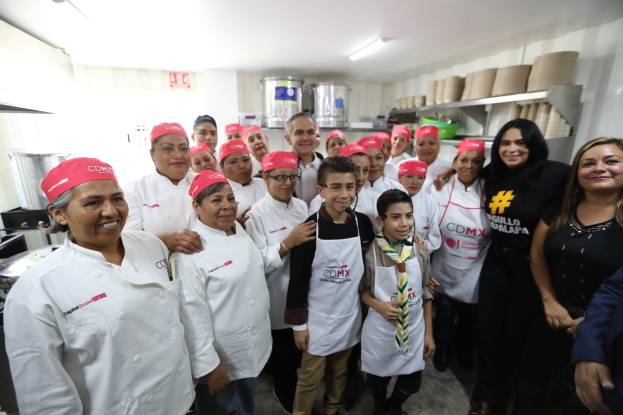 Eine Gruppe von Menschen in einer Küche, die fr├Âhlich in die Kamera l├Ąchelt und posiert, mit Mützen, Utensilien, Gestellen mit Geschirr und Deckenleuchten im Hintergrund, feiert eine Kochschule in Mexiko.
