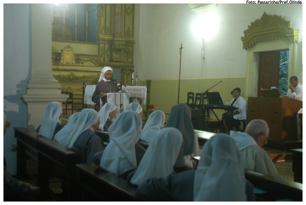 Eine Gruppe von Menschen auf einer Bank sitzend, eine Frau an einem Podium stehend, mit Stühlen, Menschen an einem anderen Podium und einer Wand im Hintergrund.