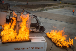 Ein Flugzeug, das auf dem Rollfeld eines Flughafens in Flammen steht, mit einem Fahrzeug und Menschen in der Nähe und Bäumen im Hintergrund.
