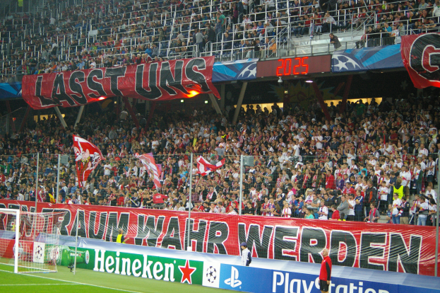 Ein Fußballspiel wird in einem Stadion mit einer großen Zuschauermenge, grünem Rasen, einem Torpfosten links, Bannern am Boden und Menschen mit Fahnen, sowie Geländern, Tafeln, Lampen und anderen Gegenständen im Hintergrund gespielt.