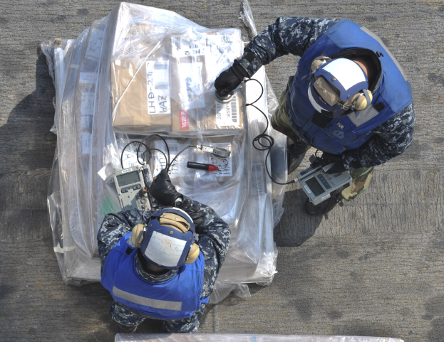 Zwei Männer in blauen Uniformen und Helmen arbeiten an einem großen Drohnen, halten verschiedene Gegenstände und haben eine Plastikabdeckung auf dem Boden.