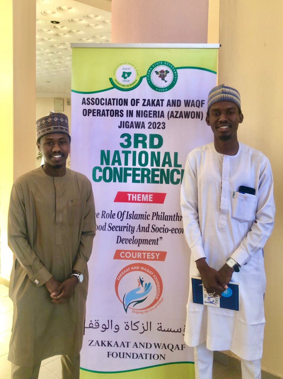 Zwei Männer neben einem Banner "3. Nationaler Kongress" mit einer Säule und Deckenbeleuchtung im Hintergrund.