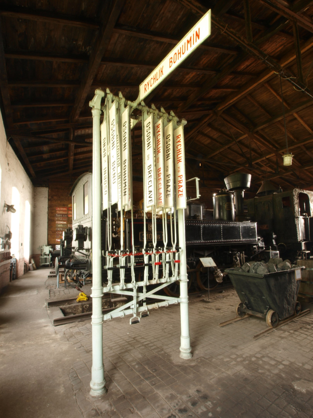 Ein Bahnhof mit einem Zug auf den Schienen, einem Wagen auf dem Boden, einem Signalkasten in der Mitte, einem Pfahl mit einer Tafel, einer Wand auf der linken Seite und einer Decke oben.