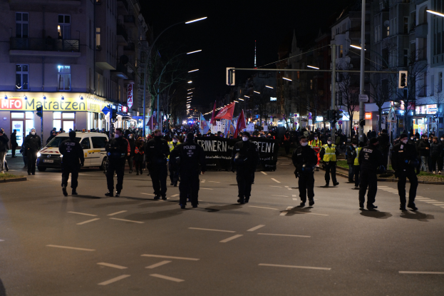 Eine Gruppe von Menschen geht nachts eine Straße in Berlin, Deutschland, während einer Demonstration, beleuchtet von Straßenlaternen und umgeben von Gebäuden, Bäumen, Laternenmasten, Verkehrszeichen und Fahrzeugen, mit einer Fahne und Schildern mit Text im Hintergrund.