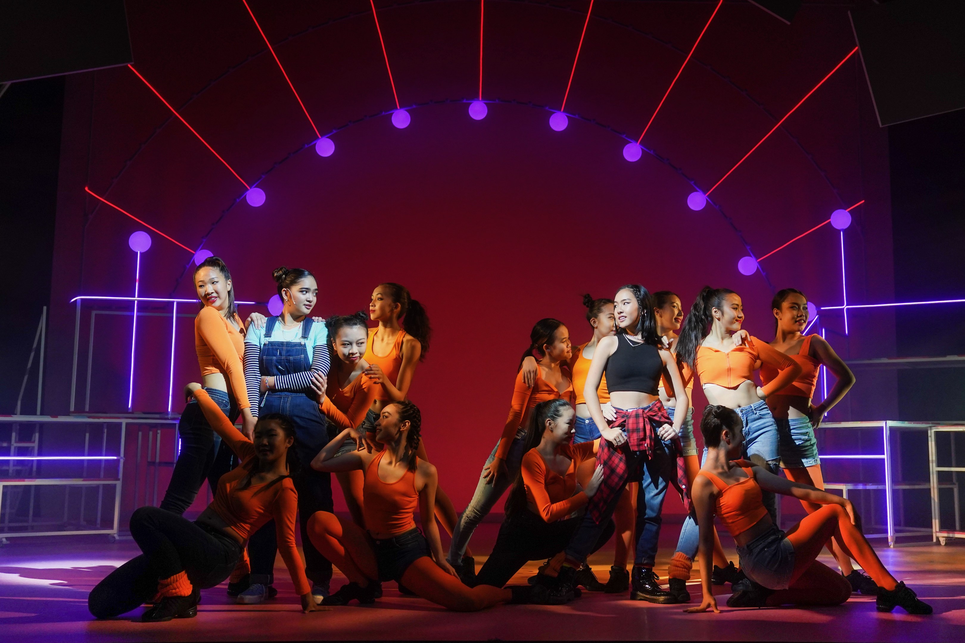 Junge Tänzer in bunten Kostümen tanzen auf einer hell erleuchteten Bühne mit gemusterten Dekorationen, Arme erhoben, während ein applaudierendes Publikum hinter Geländern zusieht.