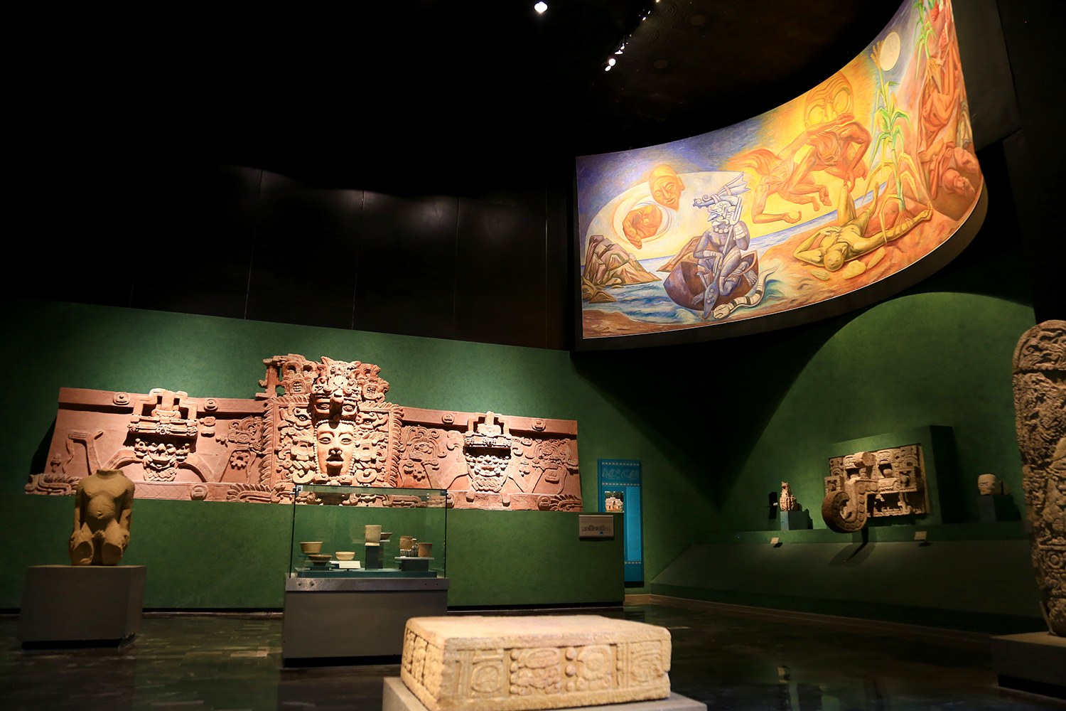 Ausstellungsraum im Anthropologischen Museum von Mexiko-Stadt, der verschiedene Kunstwerke zeigt, darunter Skulpturen, Tischobjekte, eine Texttafel, eine Wandmalerei und Deckenbeleuchtung.