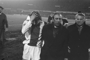 Schwarzes und weißes Bild einer Gruppe von Männern auf einem Fußballfeld mit einem Stadion im Hintergrund, die nach einem Sieg betrübt aussehen.
