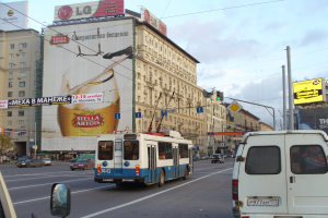 Eine belebte Stadtstraße mit mehreren Fahrzeugen, darunter ein Bus und ein Lieferwagen, umgeben von Gebäuden, Schildern, Strommasten mit Drähten und Ampeln unter einem bewölkten Himmel.