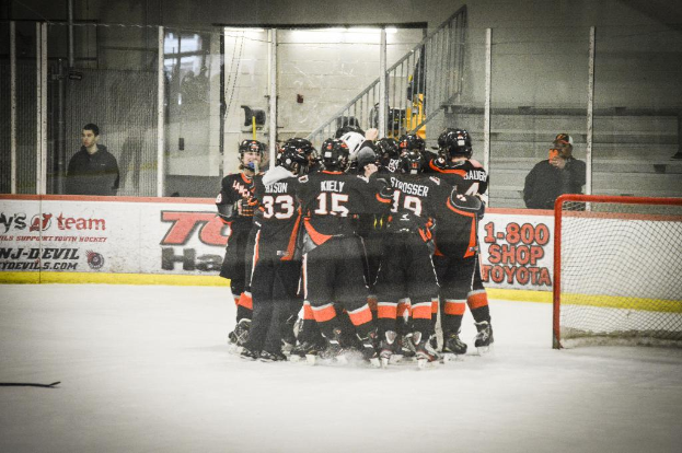 Eine Gruppe von Menschen in Schlittschuhen, Handschuhen und Helmen steht auf einem Eisboden mit Absperrungen und Glastüren dahinter, ein Hockey-Tor mit einem Netz auf der rechten Seite und ein Hockey-Schläger auf der linken Seite.